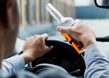 partial view of man driving car while holding bottle of whiskey, blurred foreground